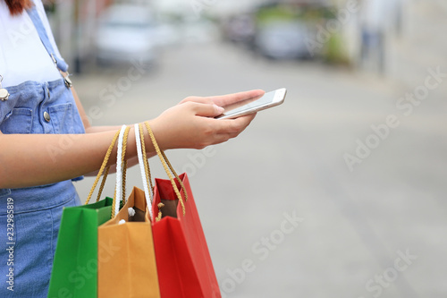 young woman hand holding smartphone and shopping bags with standing at the car parking lot