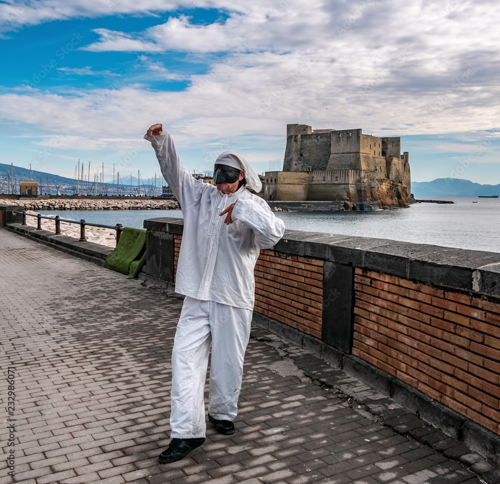 Pulcinella traditional Neapolitan mask gestures with Castel dell'Ovo in the background in the Gulf of Naples.