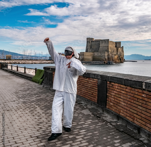 Pulcinella traditional Neapolitan mask gestures with Castel dell'Ovo in the background in the Gulf of Naples. photo