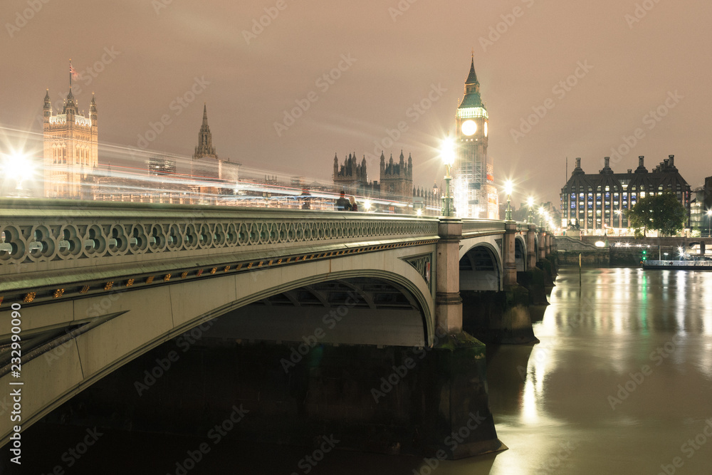 Westminster Bridge at night in the background you can see Big Ben. On the bridge you can see the blurry lights of urban transport. 