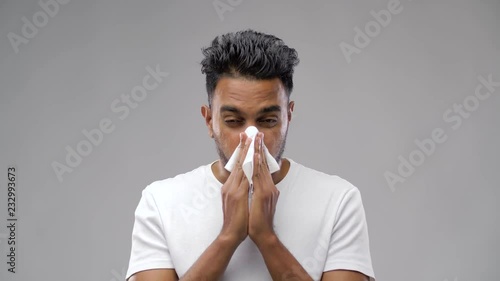 people, healthcare, rhinitis, cold and allergy concept - unhealthy indian man with paper napkin blowing nose over gray background