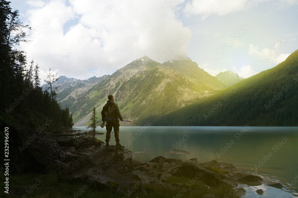 Silhouette of a male tourist against the background of a lake and mountains. A man tourist stands on the shore of a beautiful mountain lake against the backdrop of high mountains.