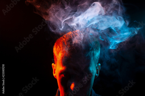 The face of a man in shadow and head is highlighted in red, who smokes a vape and gives out multi-colored smoke of blue and pink above his head, causing harm to his health. The thoughts and soul. © Aleksandr Kondratov