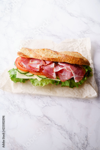 Fresh baguette sandwich bahn-mi styled. Ham, sliced cheese, tomatoes and fresh lettuce in wrapping paper on white marble background. photo