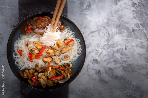 Traditional wooden chopsticks with coiled rice noodles on the background of a dish with seafood, vegetables and peppers. Copy space. Top view photo