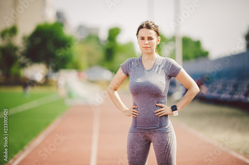 Portrait of a beautiful young caucasian woman with long hair in the tail and big breasts posing in gray sportswear standing training on a running stadium, a red rubber track in summer on a sunny day