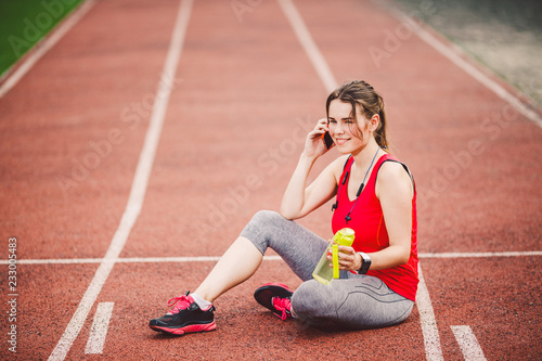 Sport and technology. A beautiful young Caucasian woman with ponytail sitting resting after workout during run at stadium, a red treadmill track. Uses for cell phone headphones and sports watches