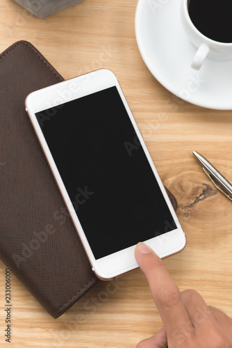 Male hand holding smartphone with notebooks, pen, and coffee cup beside on wood table in morning time