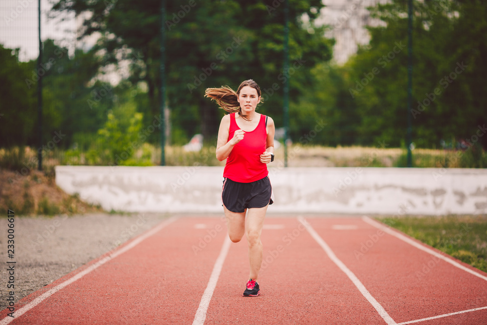Beautiful young athlete Caucasian woman with big breasts in red T-shirt and short shorts jogging, running in the stadium with red rubber coating