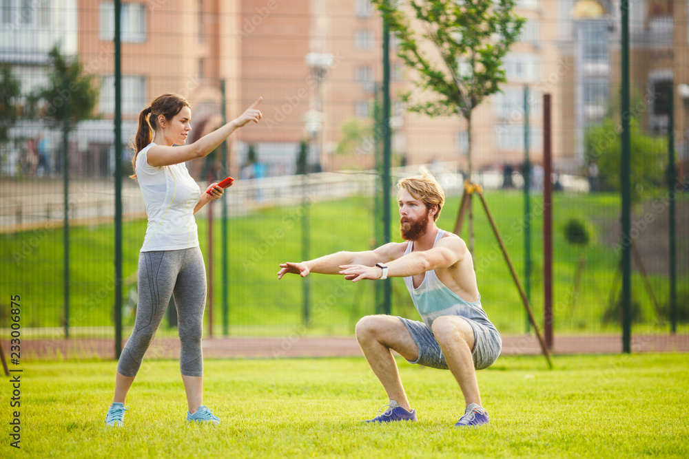 Team work in sports. A female trainer is training a young handsome man with a beard in a park on green grass, a lawn at sunset. A girl holds a phone in her hand and uses a stopwatch