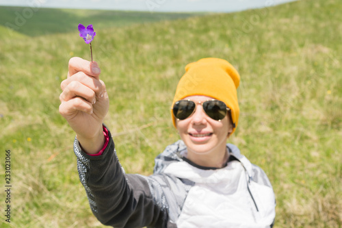 Portrait of a happy cheerful girl in a hat and sunglasses gives you a field flower against a background of green fields