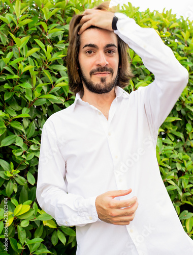 Half-length portrait of attractive young man with beard and long hair dressed in white shirt smiles at the camera while touching his hair en un parque con plantas detrás.