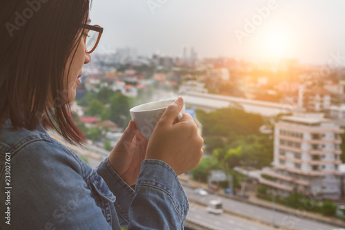 Asian woman feeling cold wearing glasses and blue denim jeans jacket holding white coffee or tea cup with softy smoke stream and looking top view foggy cityscape with sunlight radial and cloudy sky. photo
