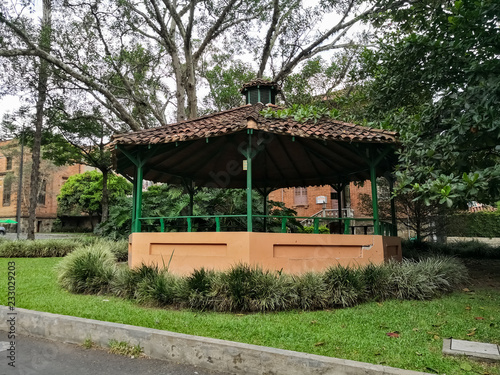 Small traditional kiosk or gazebo with green wooden columns and small circular roof with clay tiles. Green area with vegetation and trees.