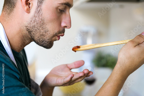 Obraz na płótnie Handsome young man tasting the fried with wooden spoon in the kitchen at home