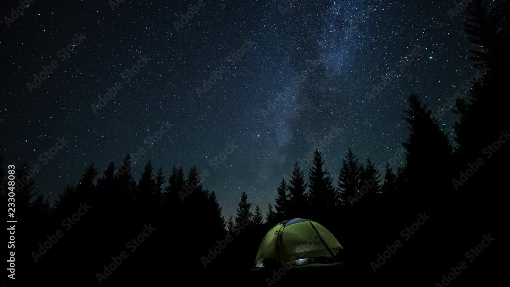 The Milky Way is moving in the night sky over the silhouettes of trees and glowing tent. Timelapse. 4K