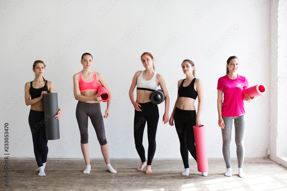 Group of young sporty woman with yoga mats standing at white wall.