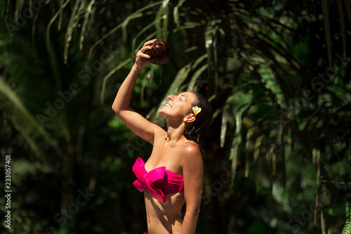 Sexy slim brunette young female watering herself with fresh coconut milk at wild green jungle background. Royal tropical resort relax