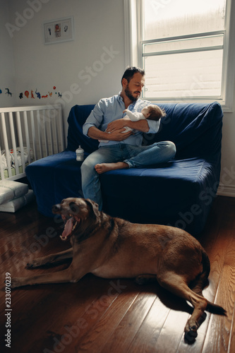 Portrait of middle age Caucasian father with newborn baby. Dog pet laying on floor. Man parent holding child in hands. Authentic lifestyle documenatry moment. Single dad family life.