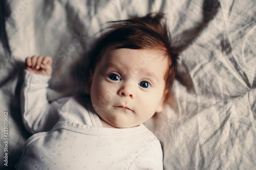 Closeup portrait of cute adorable funny white Caucasian brunette little baby newborn with blue grey eyes lying on bed looking in camera. Authentic lifestyle candid real life.Toned with film filters.