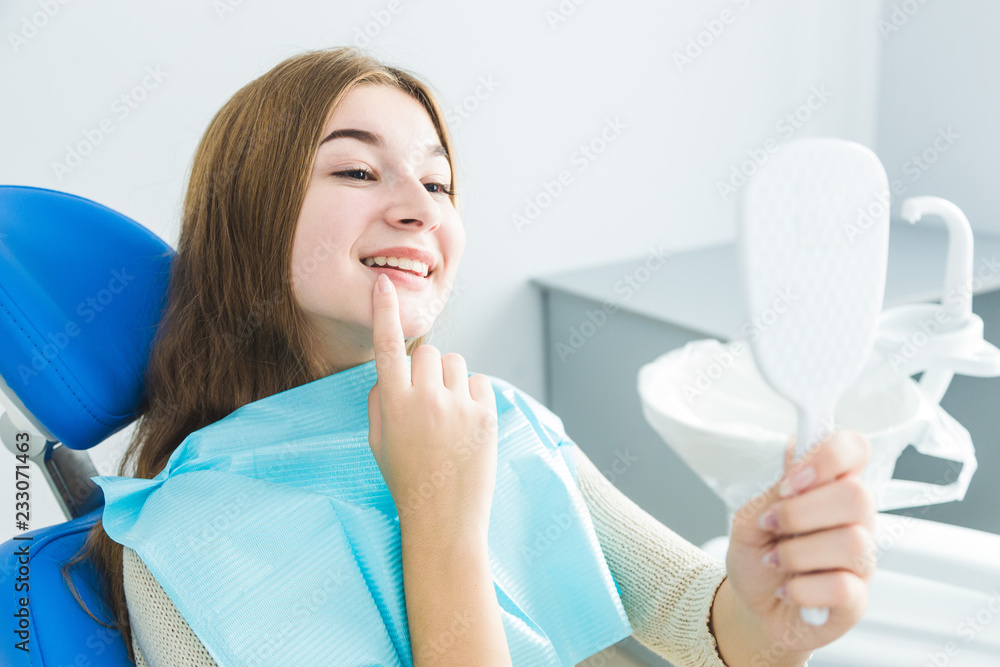 Dental clinic. Reception, examination of the patient. Teeth care. Young girl smiling, looking in the mirror after a dental checkup at her dentist Dental clinic. Reception, examination of the patient. Teeth care. Young girl smiling, looking in the mirror after a dental checkup at her dentist