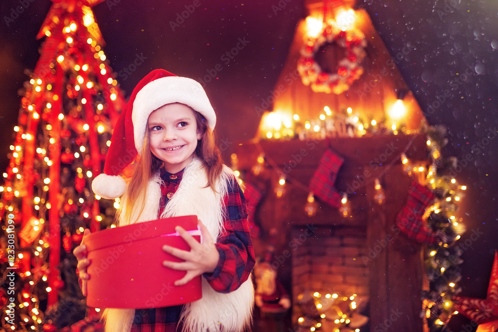 Adorable little girl in santa hat opening a magical Christmas gift by a Christmas tree in cozy living room in winter