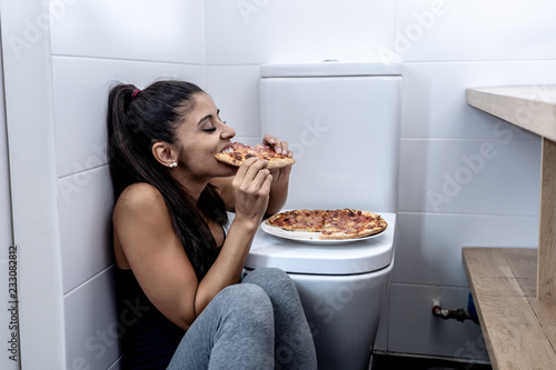 Young beautiful bulimic woman sitting on the bathroom floor eating pizza looking guilty