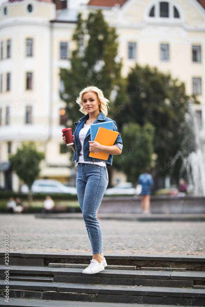 Young blonde woman student with book and coffee cup dressed in casual clothing walking around city