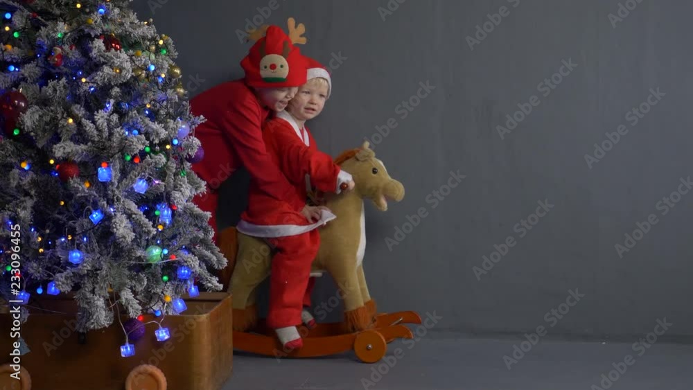 Funny children roll each other on wooden horse rocking chair on wheels in front of Christmas tree. Children are happy on holiday and are dressed in festive costumes of snowman and deer Santa Claus Funny children roll each other on wooden horse rocking chair on wheels in front of Christmas tree. Children are happy on holiday and are dressed in festive costumes of snowman and deer Santa Claus