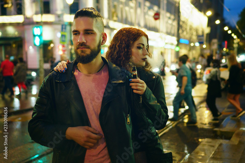 Couple in love on date. Boyfriend is ahead of her girlfriend. She hugs him and smokes an electronic cigarette. They walk streets of night city. Wet asphalt after rain.