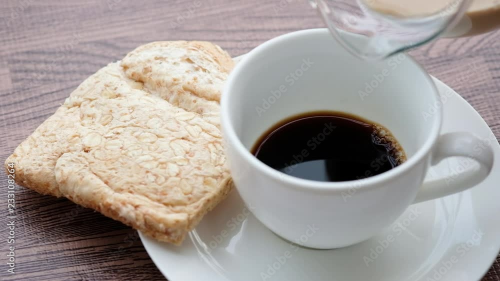 Pour fresh milk into a hot coffee glass. Breakfast cereal and Wheat bread put on a wooden table at home Pour fresh milk into a hot coffee glass. Breakfast cereal and Wheat bread put on a wooden table at home