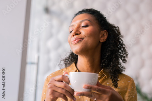Feeling joyful. Curly dark-haired appealing woman wearing yellow blouse feeling joyful while drinking coffee