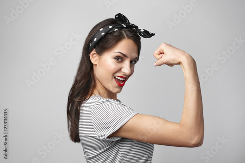 Tableau sur toile Portrait of strong independent female wearing headscarf and stylish sailor shirt showing her bicep at camera, having excited expression