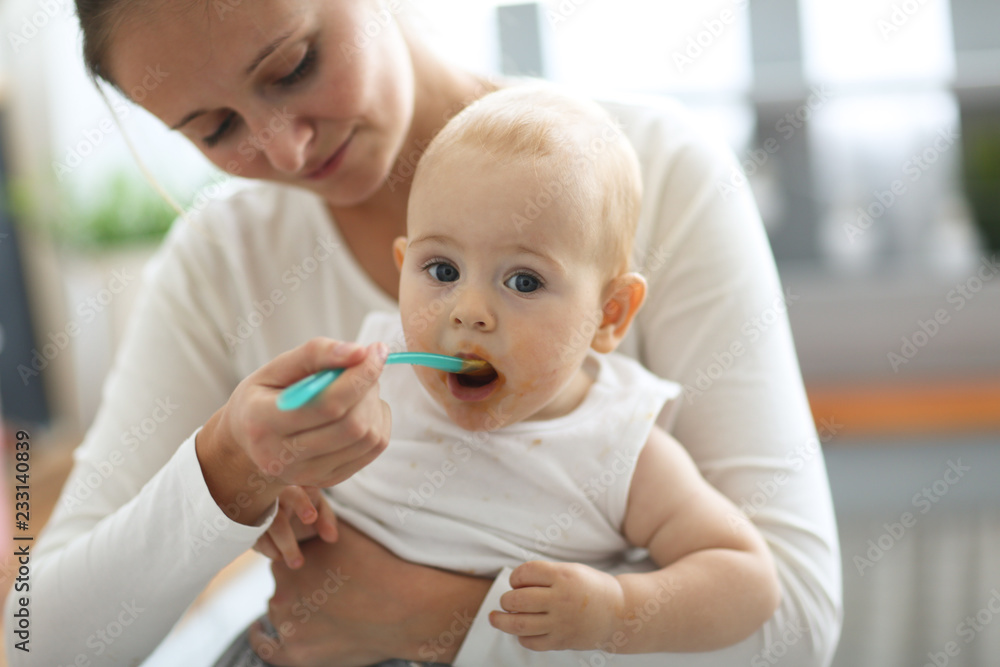 Mom feeds baby with spoon, cozy real interior Mom feeds baby with spoon, cozy real interior