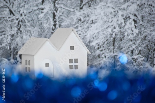Small white cardboard houses, winter snowy trees background, blue bokeh effect. Handmade home winter decoration. Do it yourself. Shallow depth of focus.