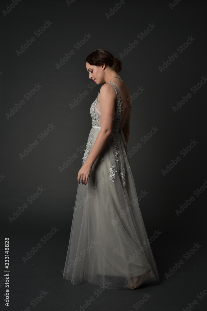 full length portrait of brunette girl wearing beautiful long gown, standing pose with background to the camera on grey studio background. full length portrait of brunette girl wearing beautiful long gown, standing pose with background to the camera on grey studio background.