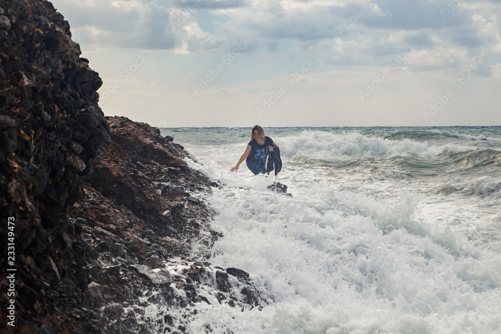 Obraz premium Woman tourist climbing on rocks at stormy sea with big waves, splashes and foam. Desperate and dangerous act