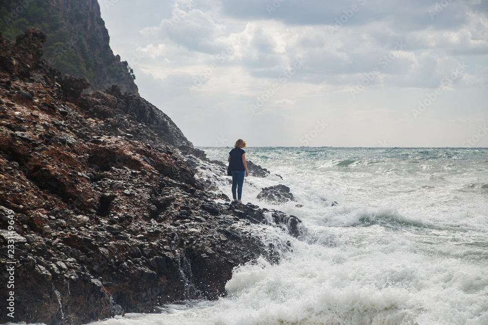 Woman tourist climbing on rocks at stormy sea with big waves, splashes and foam. Desperate and dangerous act