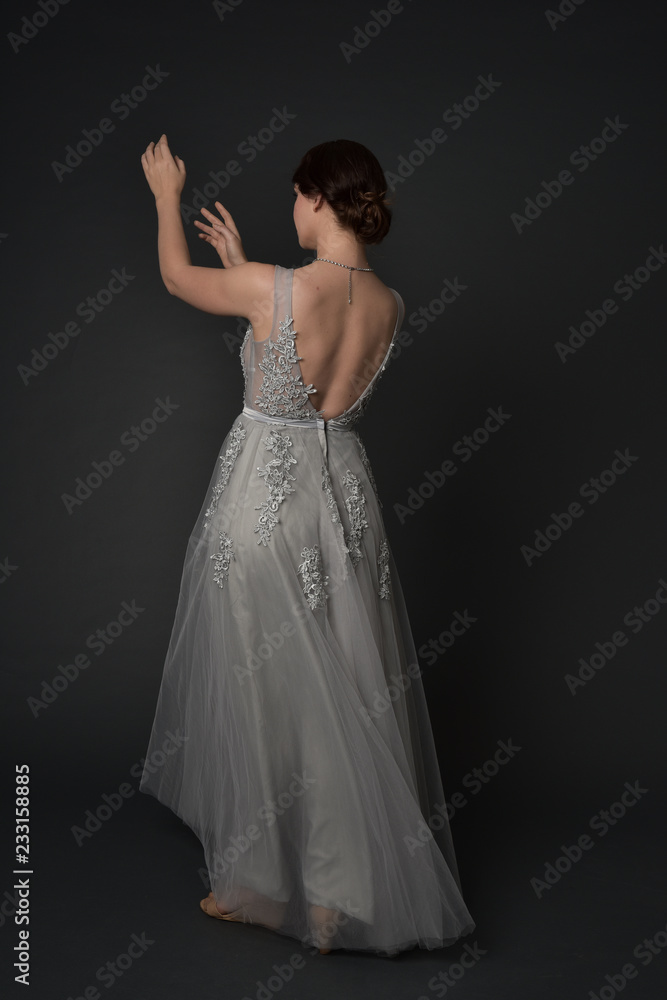 full length portrait of brunette girl wearing beautiful long gown, standing pose with background to the camera on grey studio background. full length portrait of brunette girl wearing beautiful long gown, standing pose with background to the camera on grey studio background.