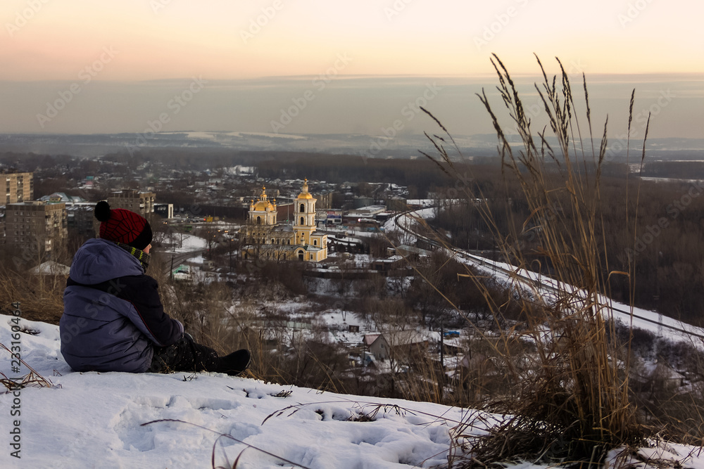 Fototapeta premium In winter, on a frosty evening in the sunset rays of the sun, the boy sits and looks at the city in front of him.