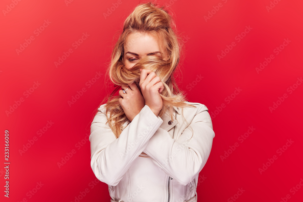 Cute smiling woman with long blonde wavy hair and in light pink jacket standing on red background and plays with her hair. Cute smiling woman with long blonde wavy hair and in light pink jacket standing on red background and plays with her hair.