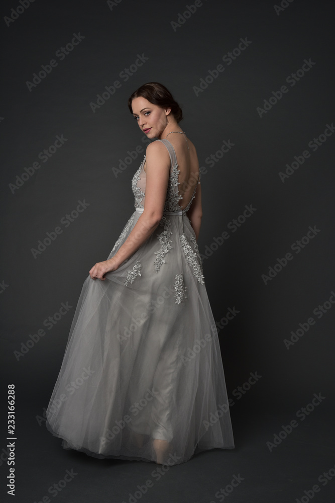 full length portrait of brunette girl wearing beautiful long gown, standing pose with background to the camera on grey studio background.