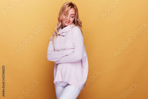 Cute woman with long blond wavy hair dressed in light pink sweater standing on yellow background with her eyes closed © monchak