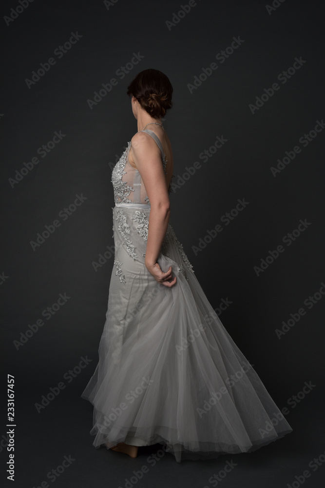 full length portrait of brunette girl wearing beautiful long gown, standing pose with background to the camera on grey studio background.