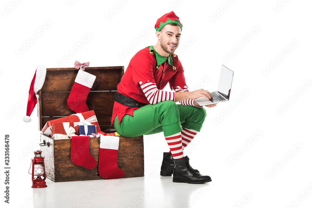 smiling man in christmas elf costume sitting on wooden box and using laptop isolated on white