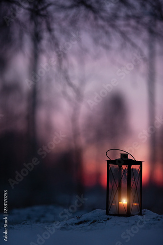 Lantern in snow against defocused sunset forest background.