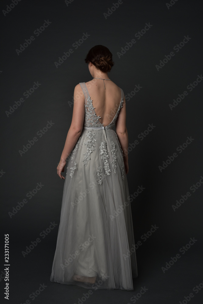 full length portrait of brunette girl wearing beautiful long gown, standing pose with background to the camera on grey studio background.