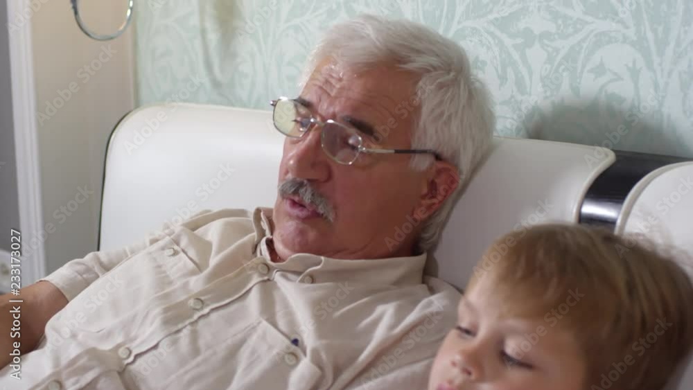 Tilt up of senior grandfather in eyeglasses lying on bed and telling something to little grandson while watching TV together at home Tilt up of senior grandfather in eyeglasses lying on bed and telling something to little grandson while watching TV together at home