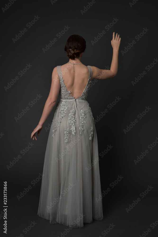 full length portrait of brunette girl wearing beautiful long gown, standing pose with background to the camera on grey studio background. full length portrait of brunette girl wearing beautiful long gown, standing pose with background to the camera on grey studio background.