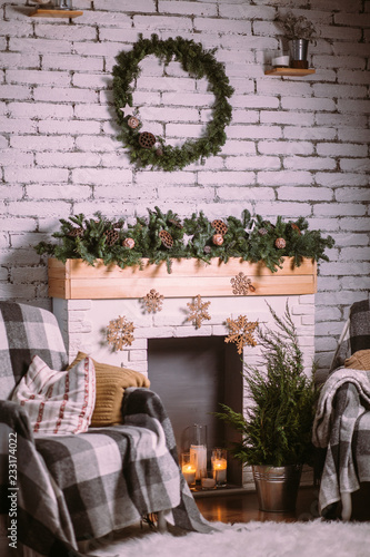 Decorative fireplace decorated with New Year's garland and burning candles in glass cups. Round green wreath hanging on white brick wall. Cozy Christmas atmosphere.